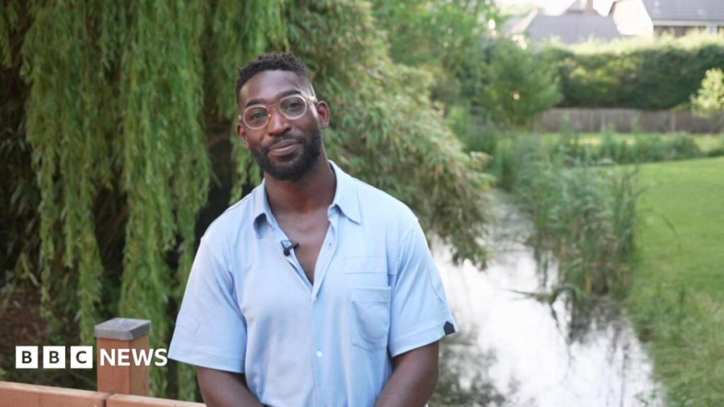 Tinie Tempah in a blue shirt and glasses smiling at the camera while on a bridge over a river in a green area.