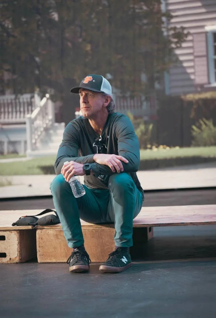 Man seated on a wooden platform, wearing a casual outfit with a baseball cap and sneakers, holding a water bottle, outdoors in a relaxed setting