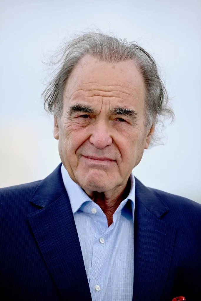 An older man in a suit, gazing at the camera with a neutral expression, standing against a plain background