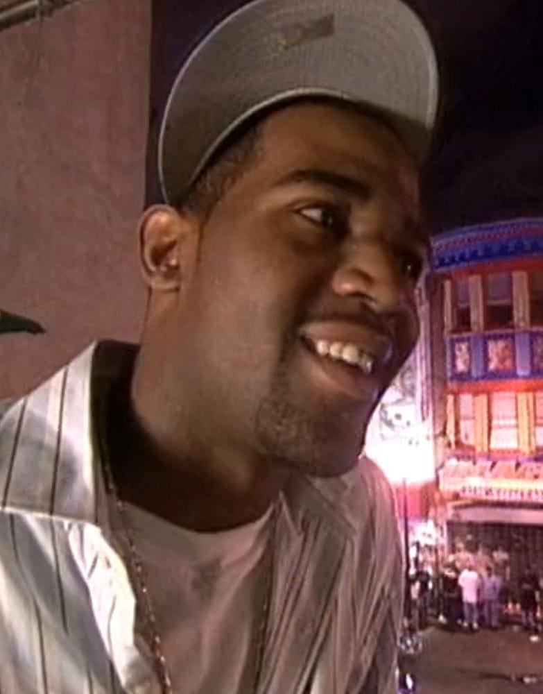 Smiling person wearing a hat and shiny shirt in an urban night scene.