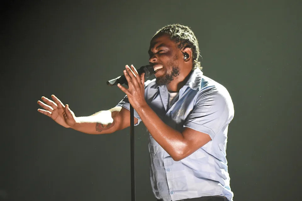 Kendrick Lamar passionately singing into a microphone on stage, wearing a short-sleeve shirt