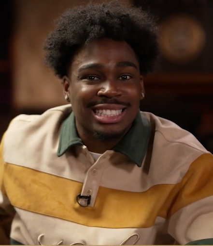 A young man with curly hair and a mustache smiles, wearing a cream and gold striped polo shirt.