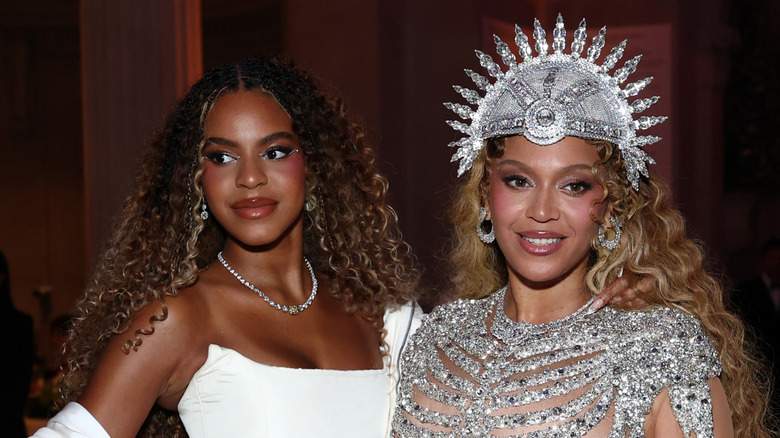 Beyoncé wearing an ornate crystal outfit and headdress standing next to daughter Blue Ivy wearing a white gown at the Met Gala.