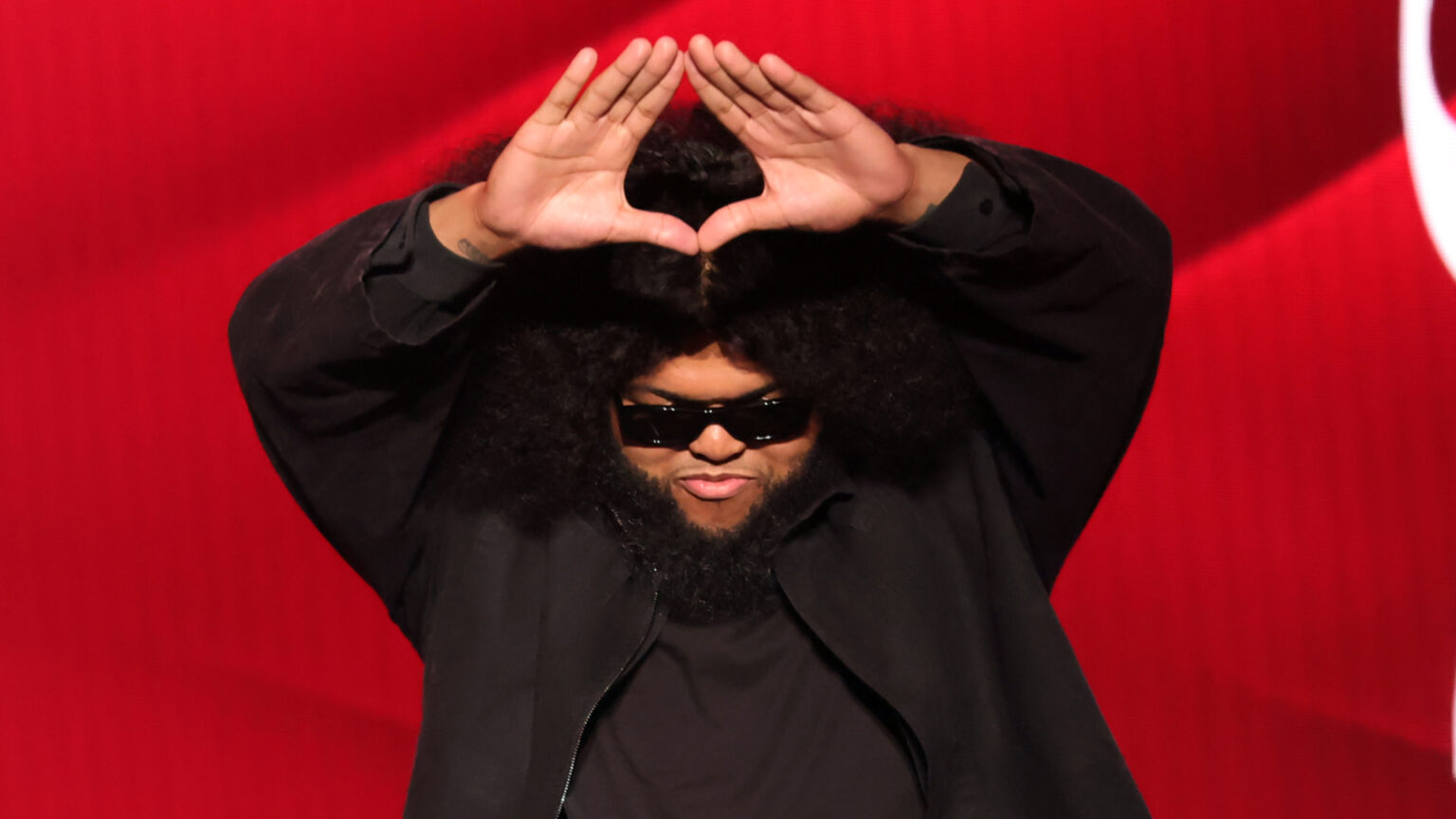 Druski makes a triangle shape with his hands against a red background, parodying Jay-Z.