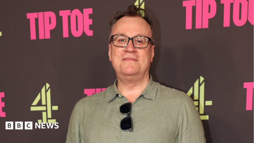 Russell T Davies reveals he is planning new project after Tip Toe Russell T Davies stands on a red carpet. He wears glasses and short dark hair, and wears a khaki green shirt with sunglasses tucked in the collar.
