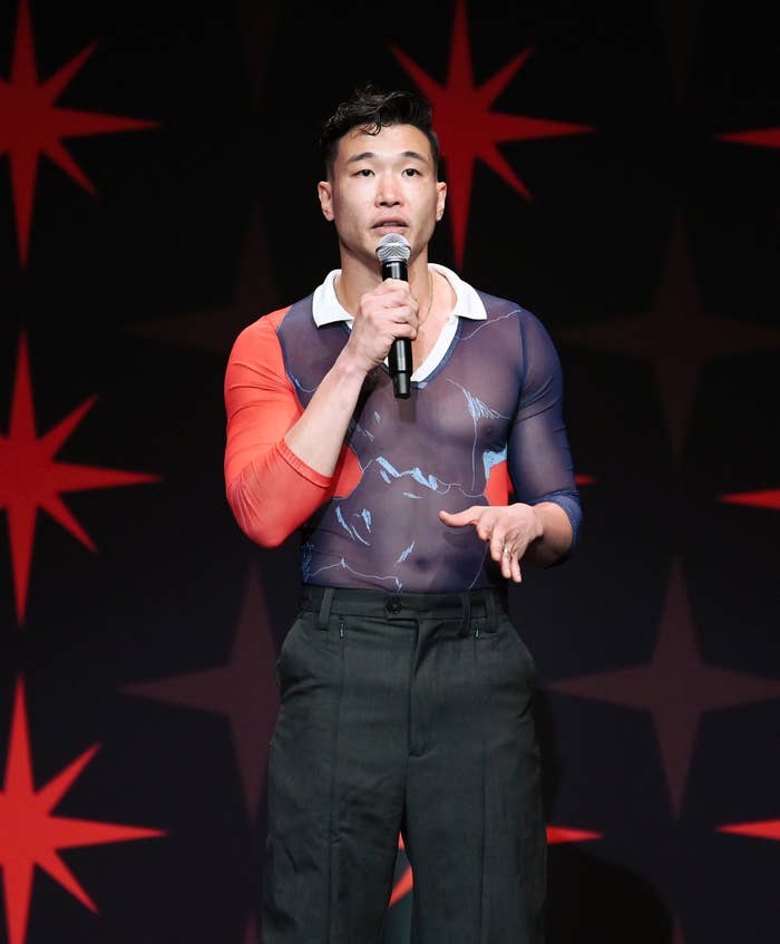 Man in a sheer top with comic motifs and black trousers speaks into a microphone on stage decorated with star patterns