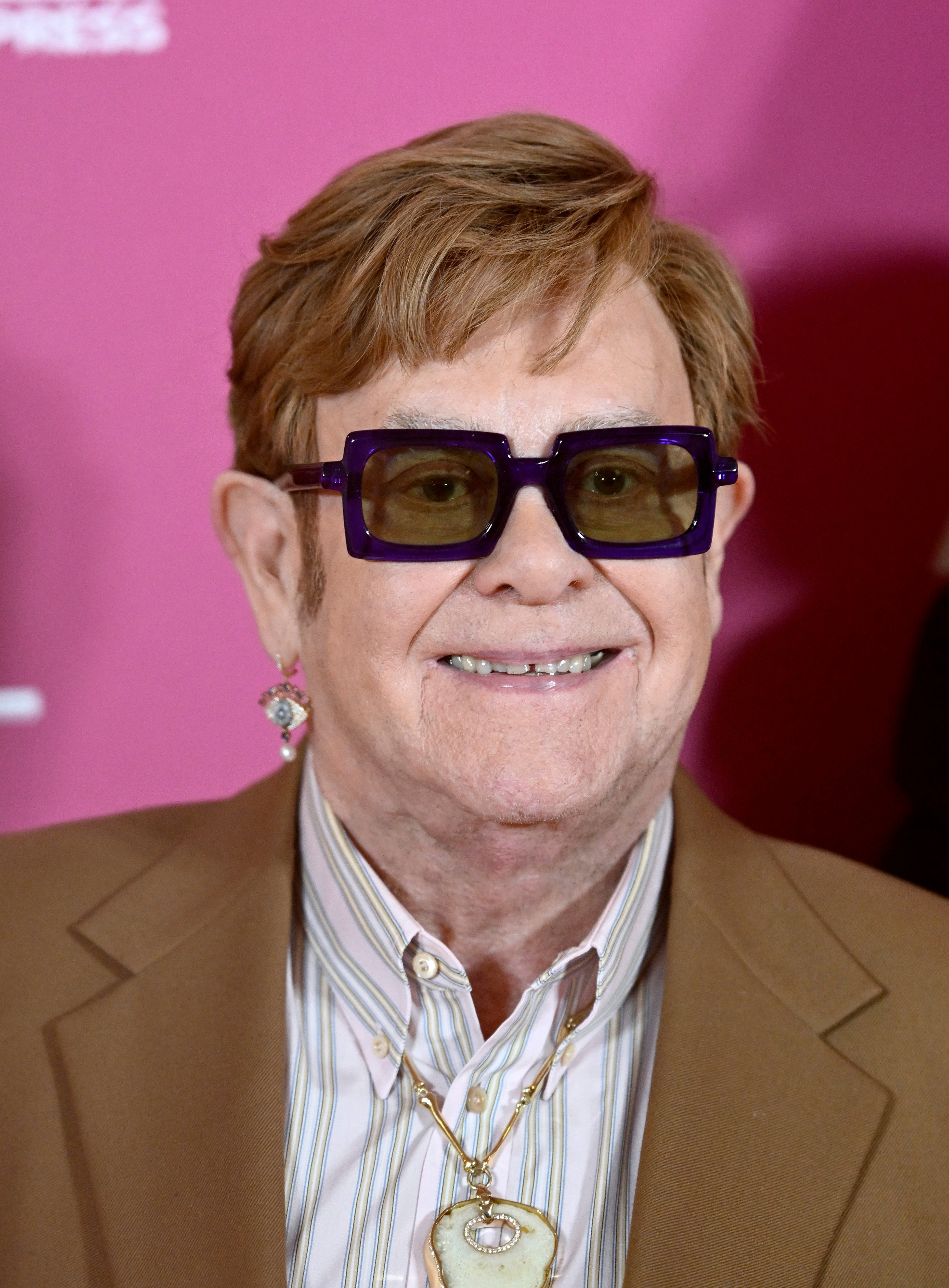 Person wearing a tan suit and striped shirt, with dark glasses and a pendant. Smiling at a formal event