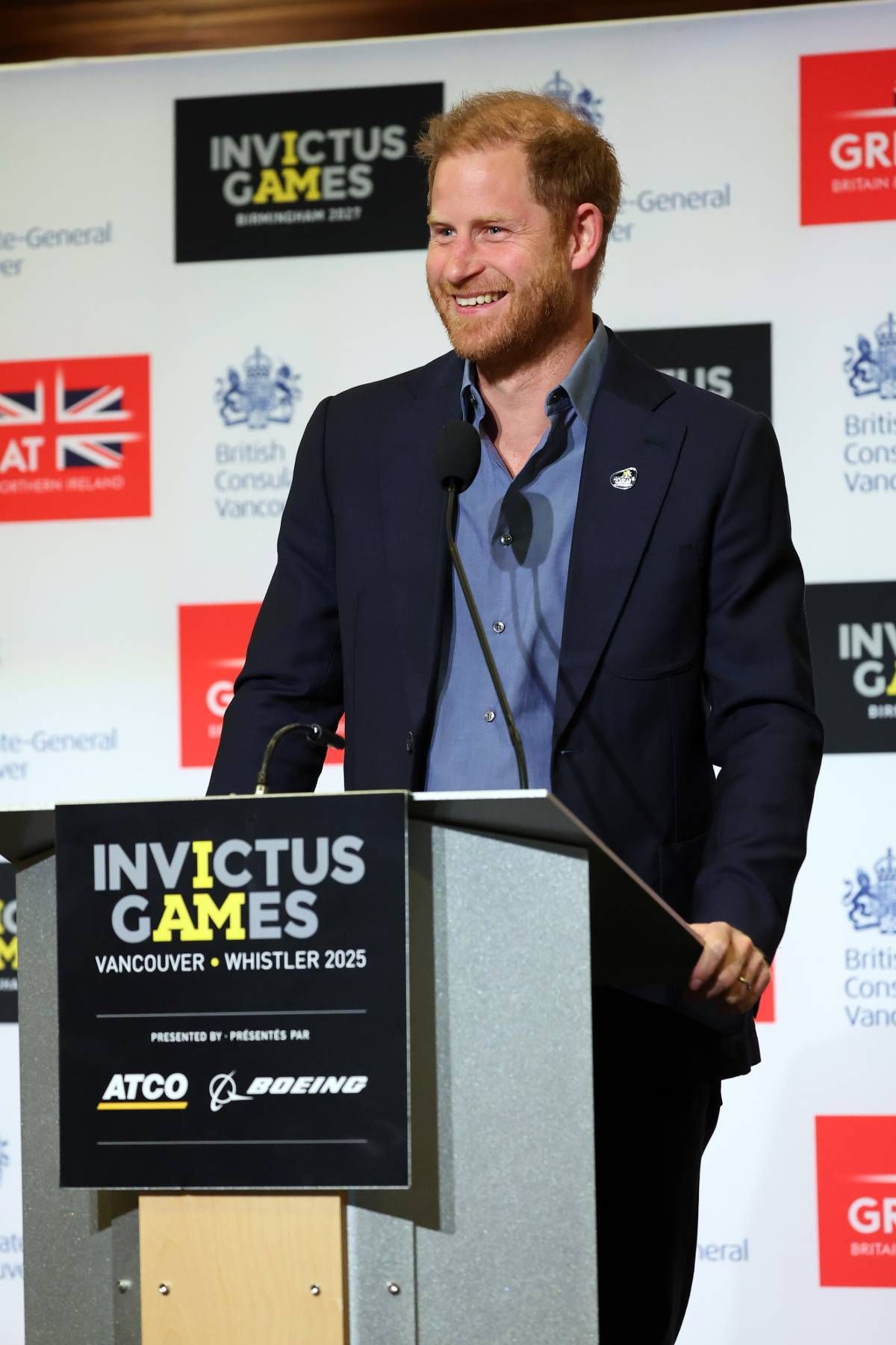 An adult male is standing at a podium, smiling and addressing an audience. Behind him, a backdrop features various logos, including "INVICTUS GAMES" and "GR," with other logos visible in the background. The individual is dressed formally in a suit and a light-colored shirt.