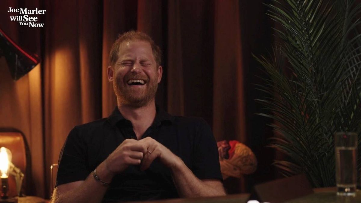 An individual, dressed in a dark-colored polo shirt, is seated with a joyful expression, their hands clasped together in front of them. The setting appears to be a cozy indoor environment, with a warm, ambient lighting and a partially visible backdrop of a curtain and decorative elements.