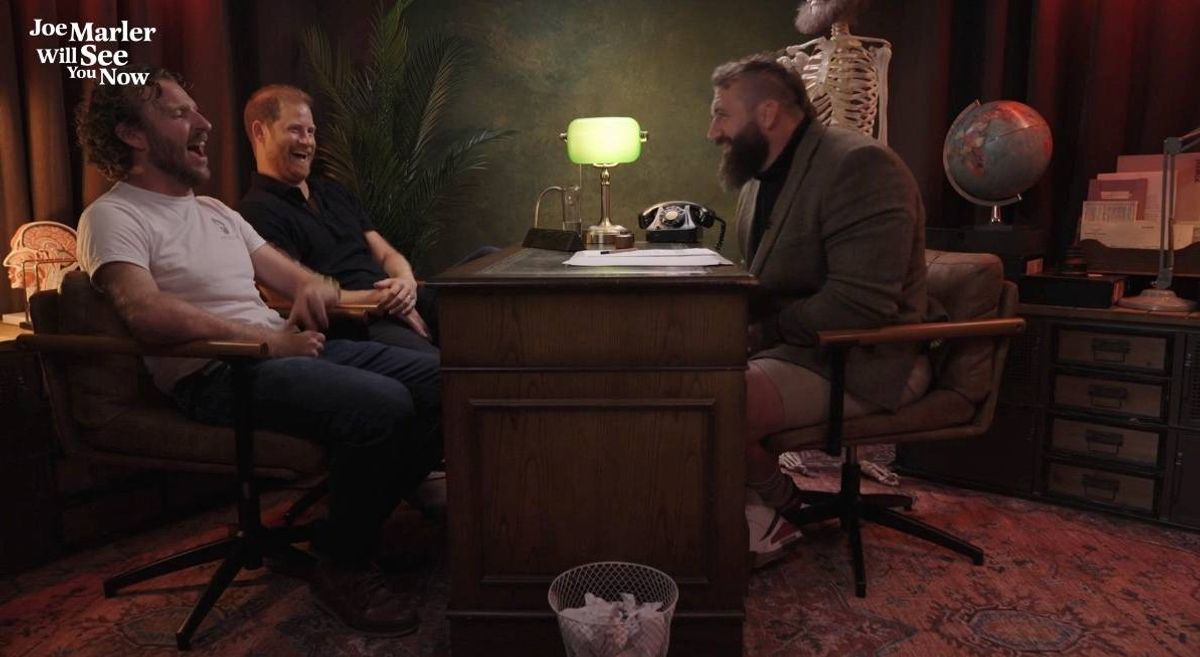 Three individuals are seated in a room, likely engaged in a conversation. A person on the left is wearing a white t-shirt and blue jeans, while the person in the middle is wearing a black t-shirt. The person on the right is dressed in a dark suit. A desk with a lamp and some papers is situated in front of them, and a trash can is placed beside the desk.