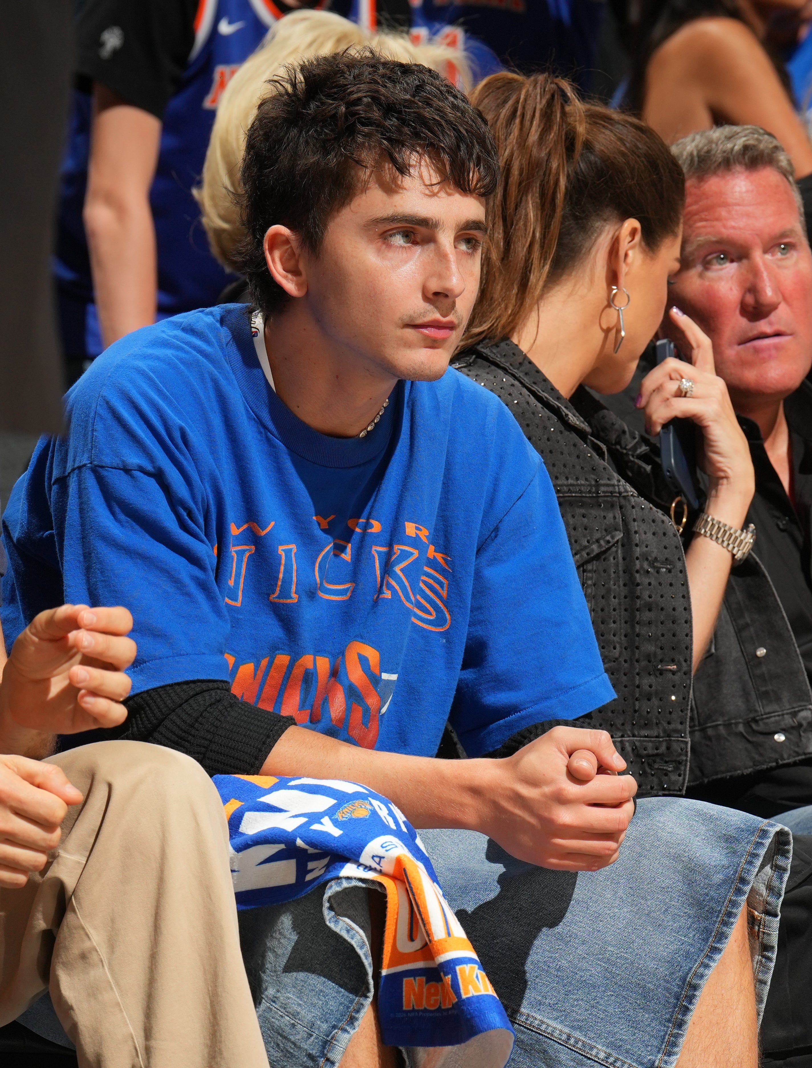 A person in a blue sports jersey sits in a crowd at an event, looking to the side, surrounded by other attendees