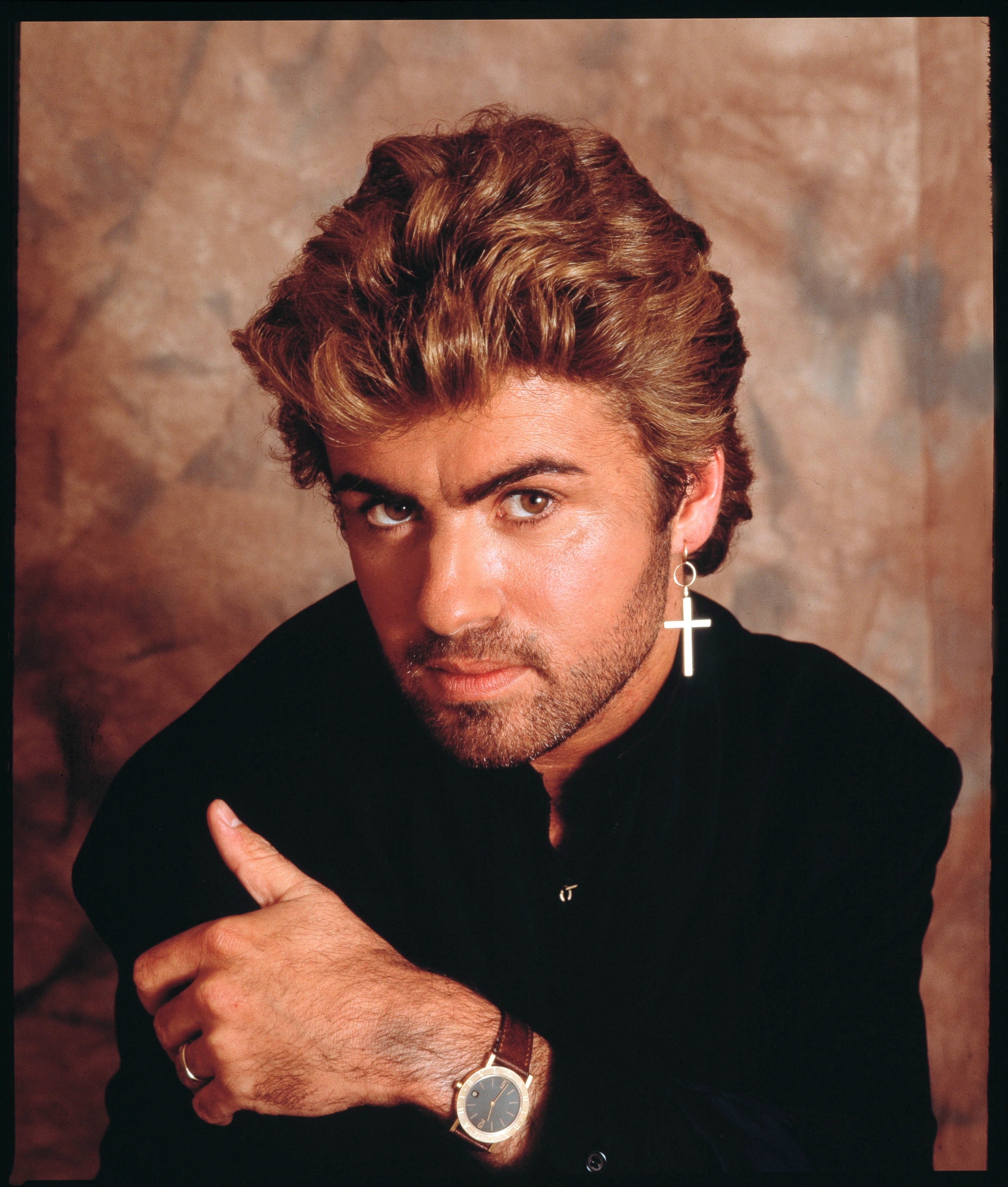 A person with styled hair, wearing a cross earring and a watch, poses confidently against a neutral backdrop