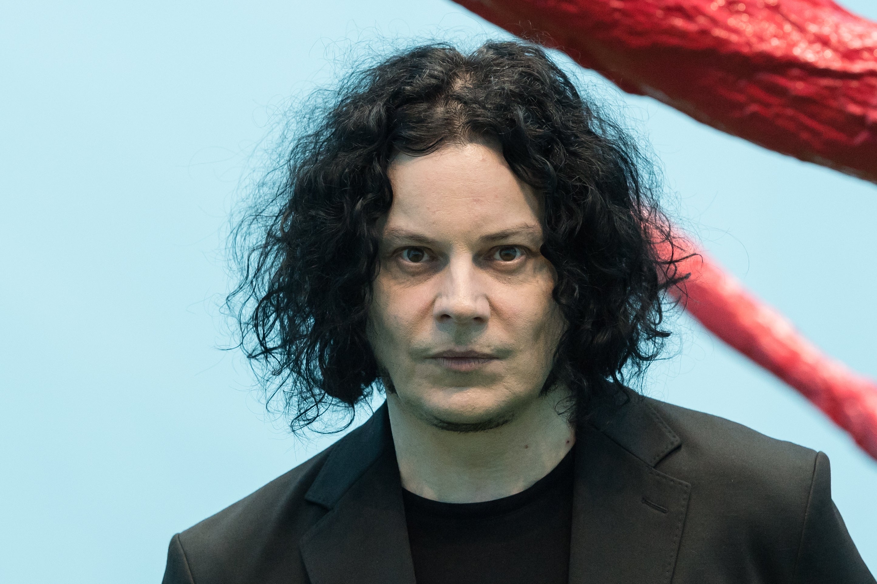 Jack White with medium-length curly hair, wearing a suit, standing in front of an abstract sculpture