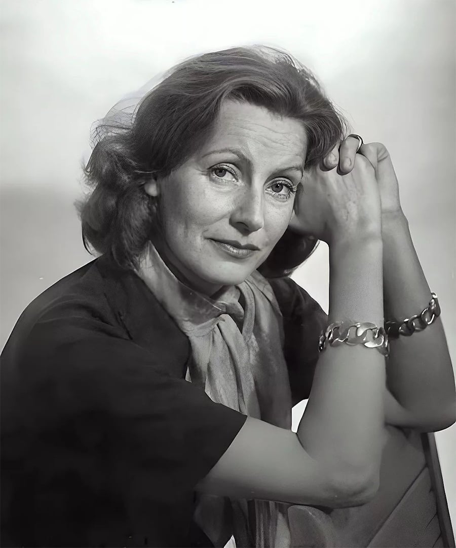 Black-and-white portrait of a woman with wavy hair, wearing a dark top, a light scarf, and a chunky bracelet, resting her arms on a chair and looking thoughtfully at the camera.