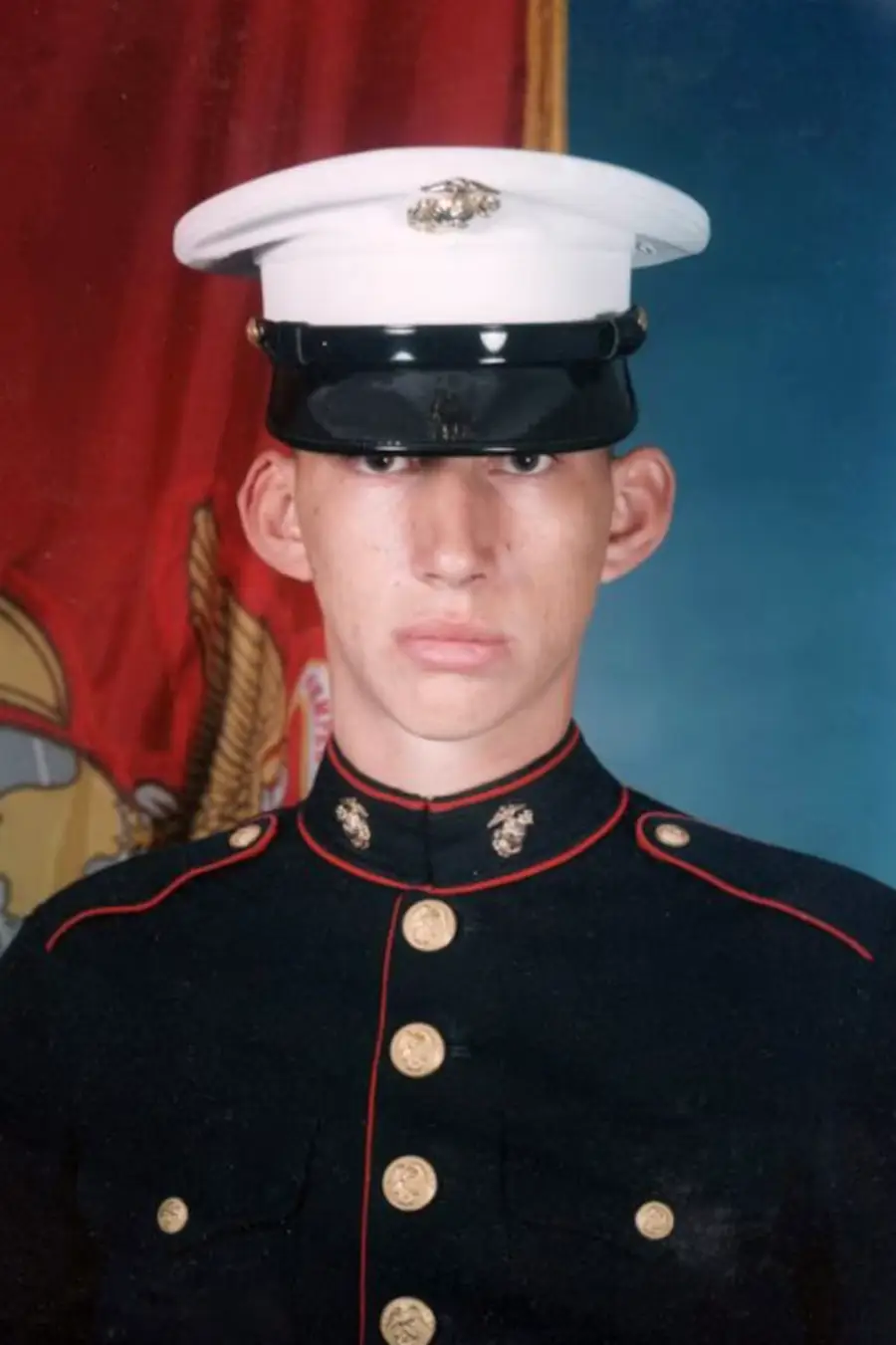 A young person in a U.S. Marine Corps dress uniform poses in front of a red flag with an emblem, looking directly at the camera with a serious expression.