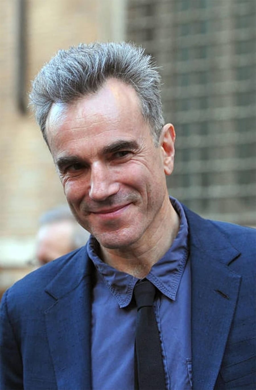 A man with short gray hair in a blue shirt, black tie, and blue jacket smiles slightly while standing outdoors with a blurred brick wall background.