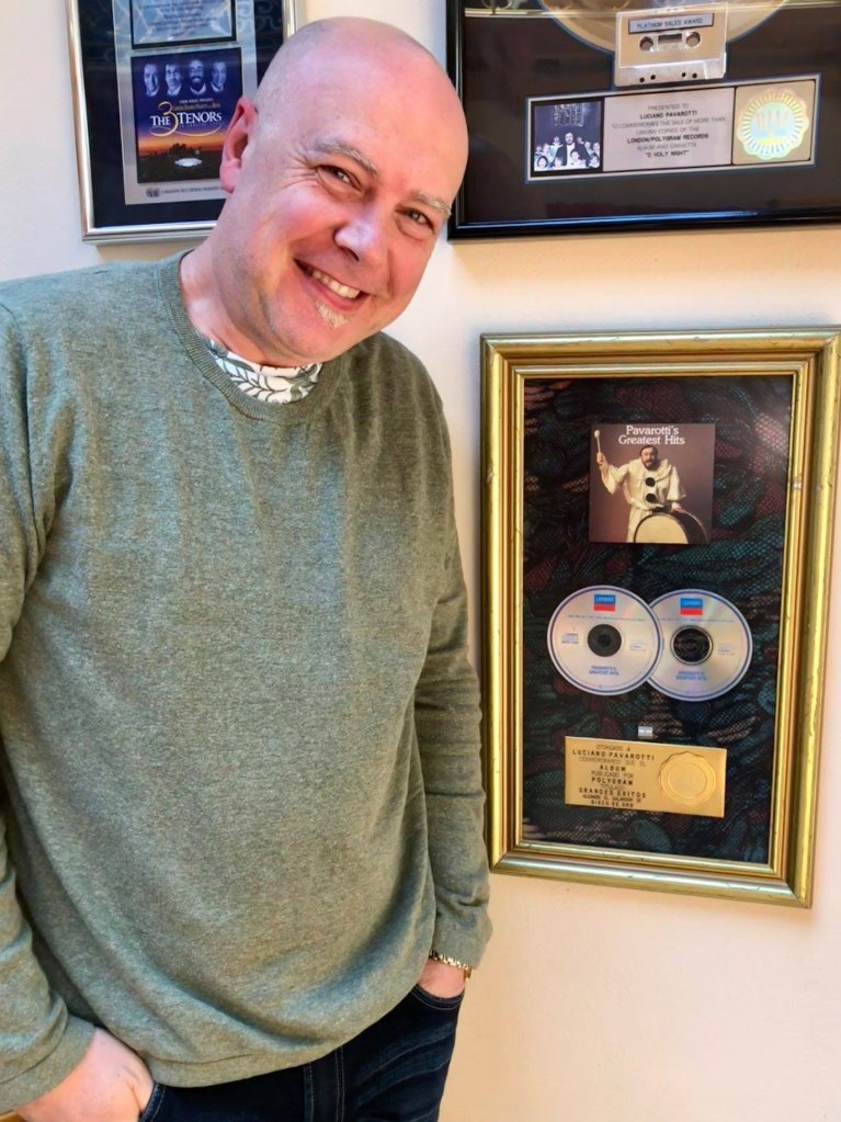 Brian Doerksen smiling next to a framed Pavarotti's Greatest Hits album, two CDs, and an inscription that reads "OTORGADO A LUCIANO PAVAROTTI CONMEMORANDO QUE EL ALBUM PUBLICADO POR POLYGRAM TITULADO GRANDES EXITOS DISCO DE ORO".