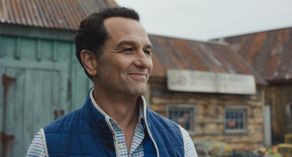 A smiling man in a blue vest standing in front of weather-worn buildings.