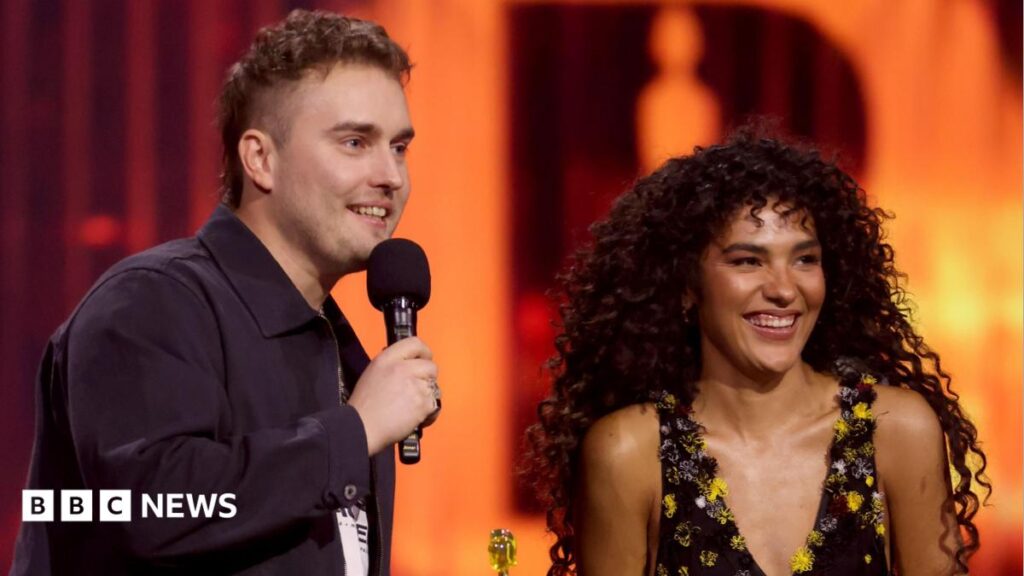 Sam Fender and Olivia Dean make chart history with Rein Me In Sam Fender and Olivia Dean speaking and smiling on stage at the Brit Awards
