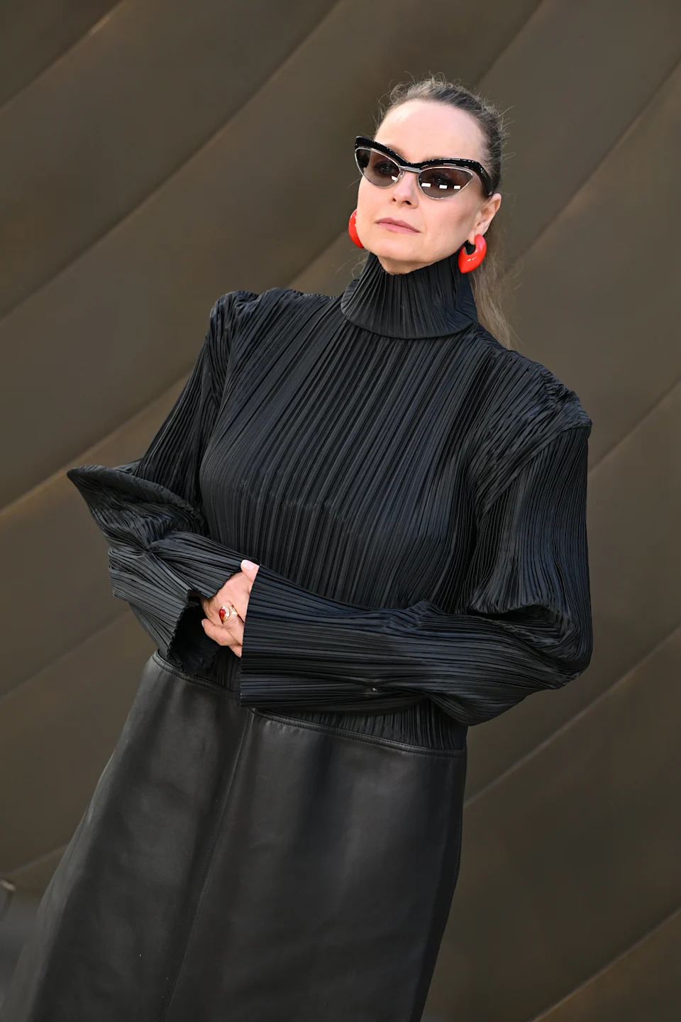 Person in pleated turtleneck dress with statement earrings and cat-eye sunglasses on a red carpet
