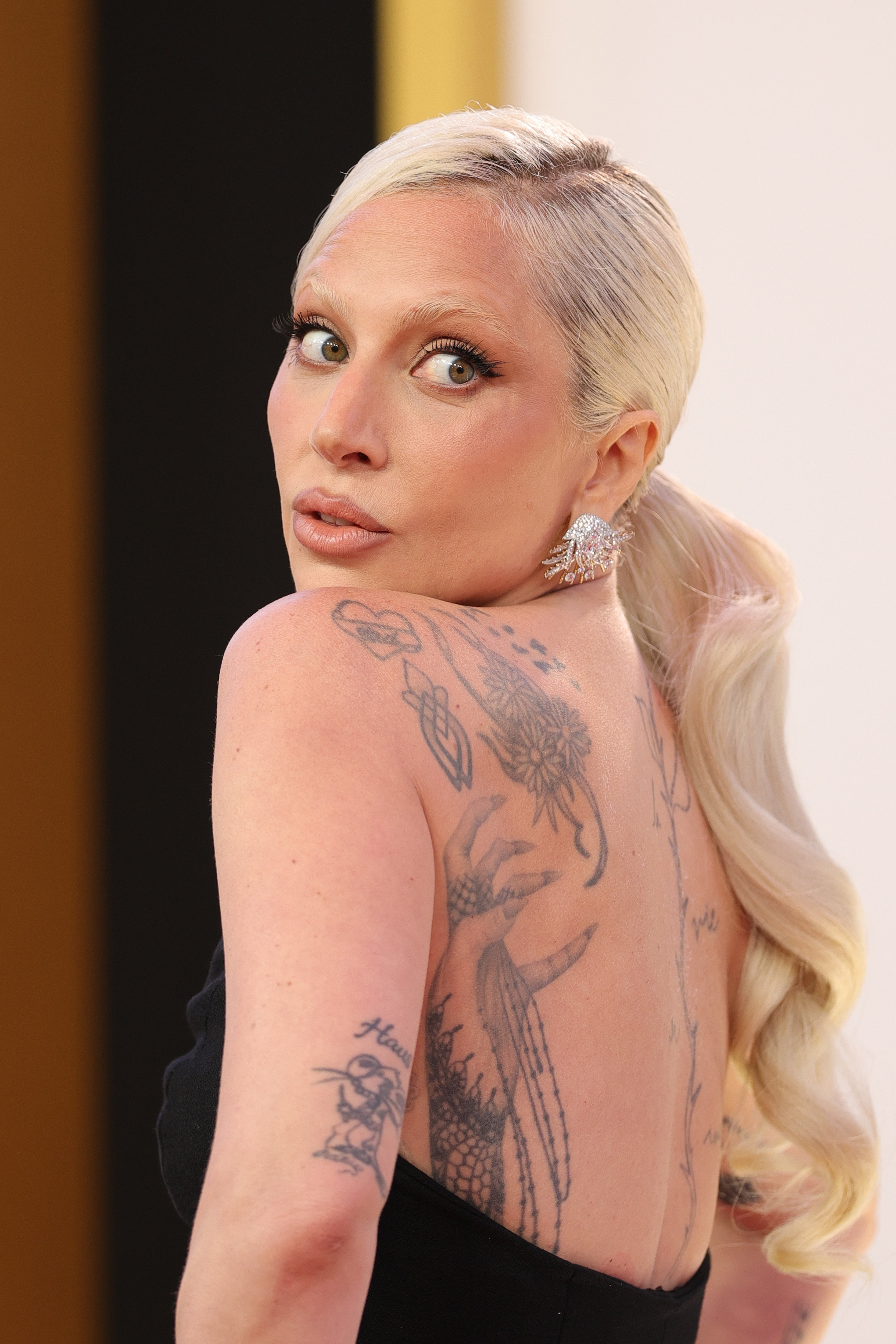 Person with tattoos and styled hair looks over shoulder, wearing a strapless gown and statement earrings