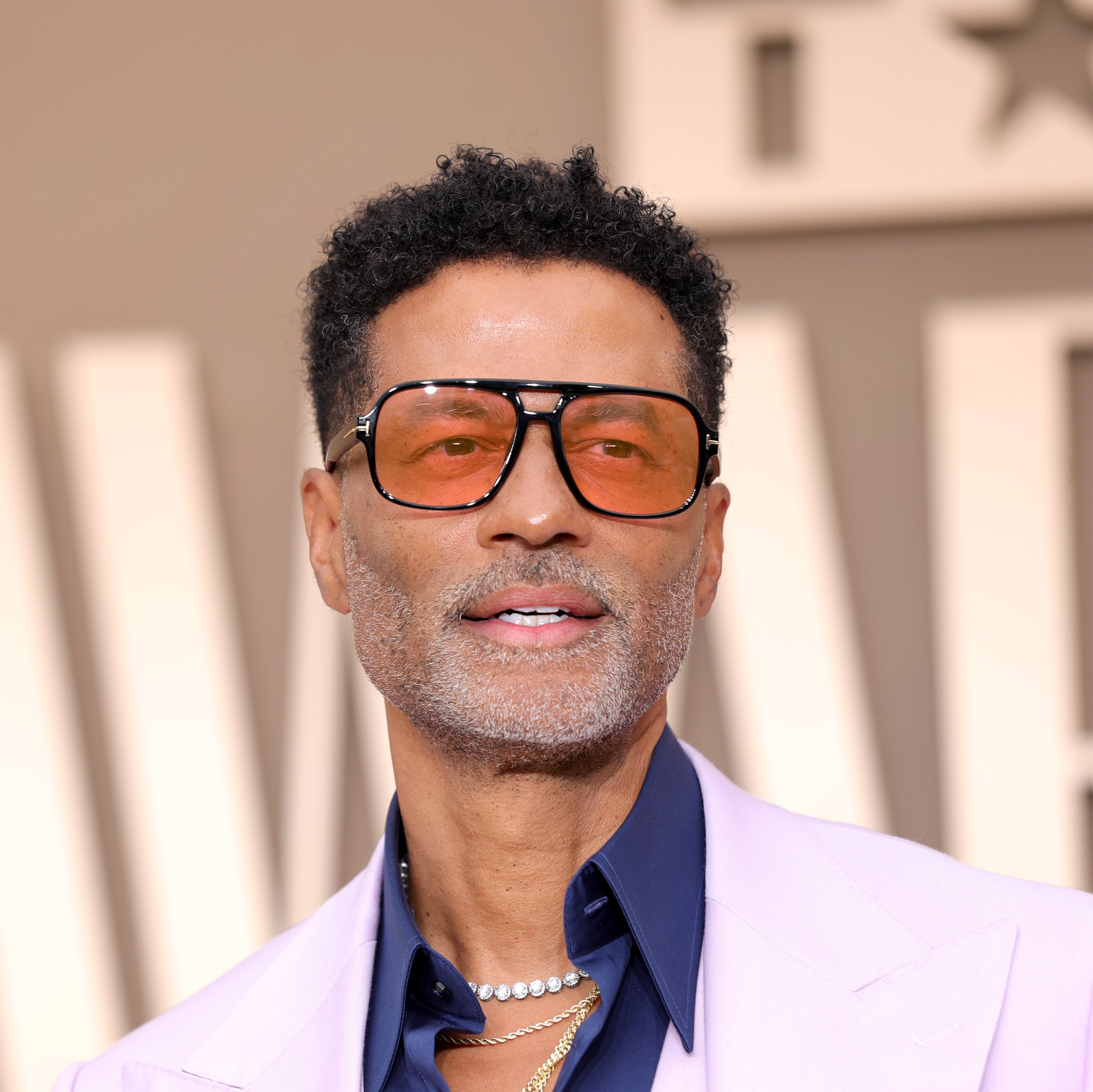 Eric Benet  in sunglasses and a light suit at an awards event, wearing a navy shirt and pearl necklaces. Simple background with large letters behind