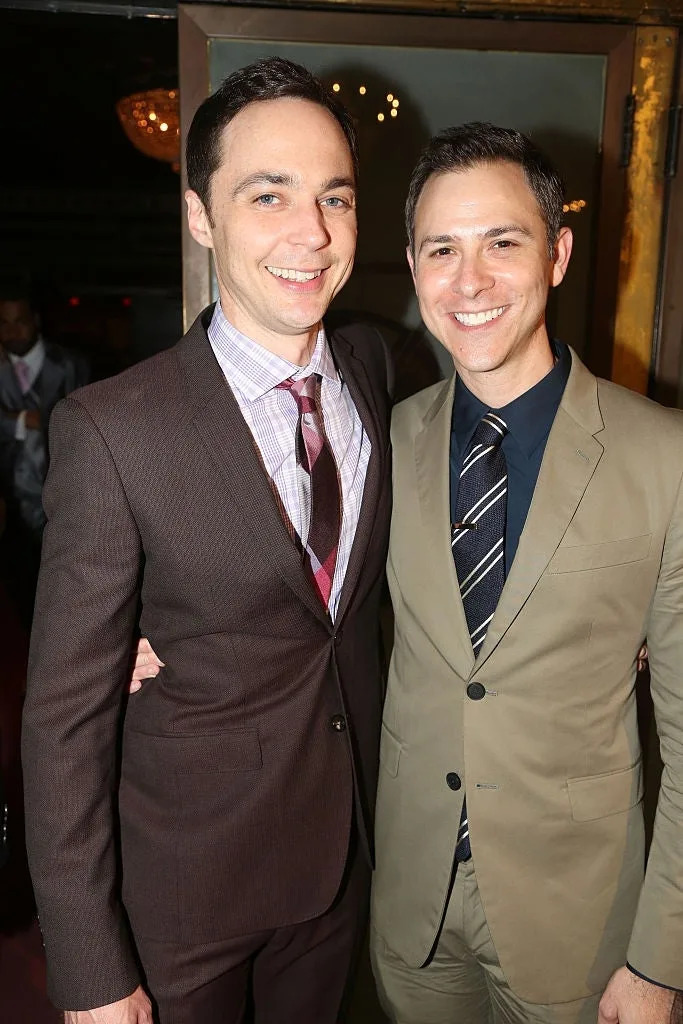 Two men smiling, one in a dark suit with a striped tie, the other in a light suit with a tie. They stand close, arm around each other, indoors