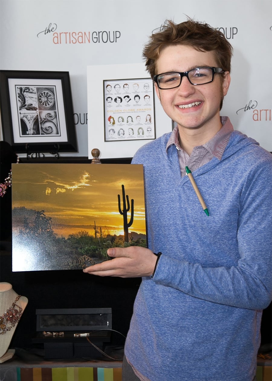 A smiling young person wearing glasses and a blue hoodie holds a framed photo of a desert sunset with a cactus. Art and jewelry are displayed on a table and wall behind them at an art event.