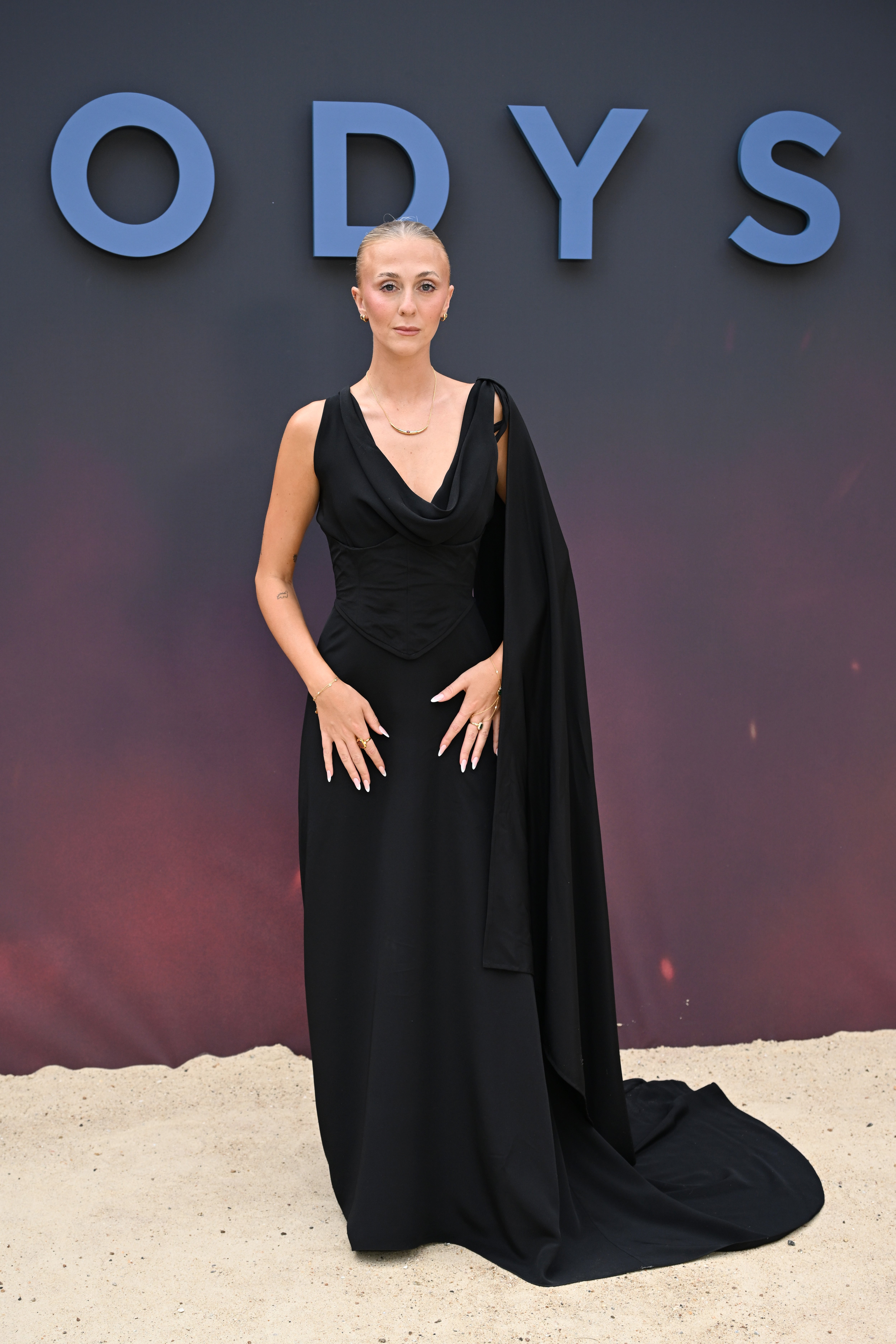 Person in elegant, flowing black gown with draped sash stands on sand against a backdrop with large letters "ODYS" visible