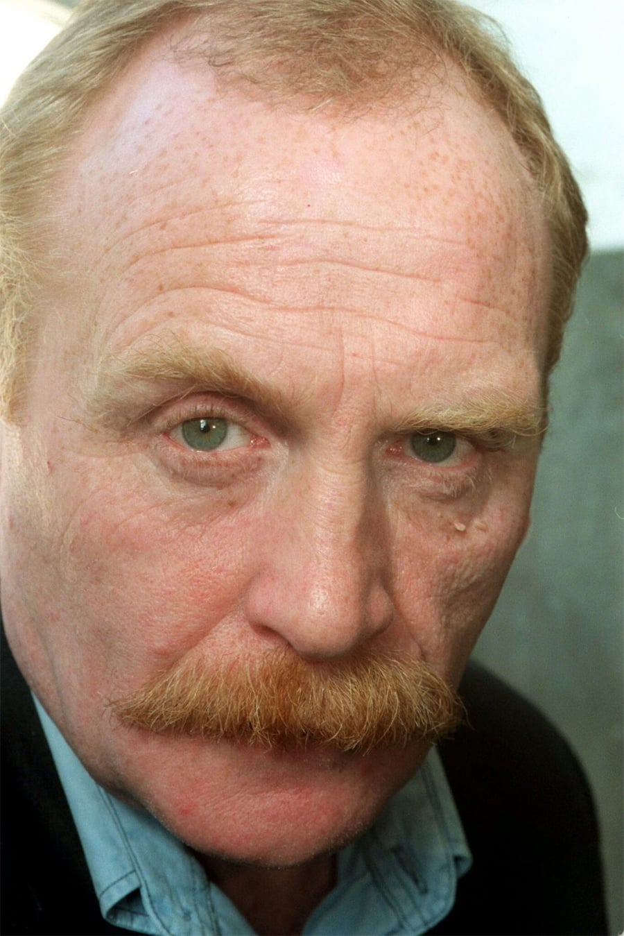 A close-up of an older man with light skin, reddish hair, a large mustache, and blue-green eyes. He is wearing a light blue shirt and a dark jacket, looking directly at the camera with a serious expression.