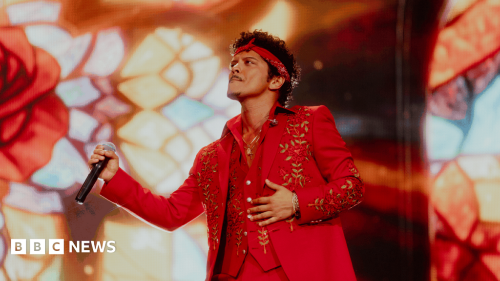Bruno Mars brings hits, romance and a Paddington Bear to Wembley Bruno Mars holds a microphone and performs at Wembley stadium wearing a red suit with gold flower detail. He also wears a red headband.
