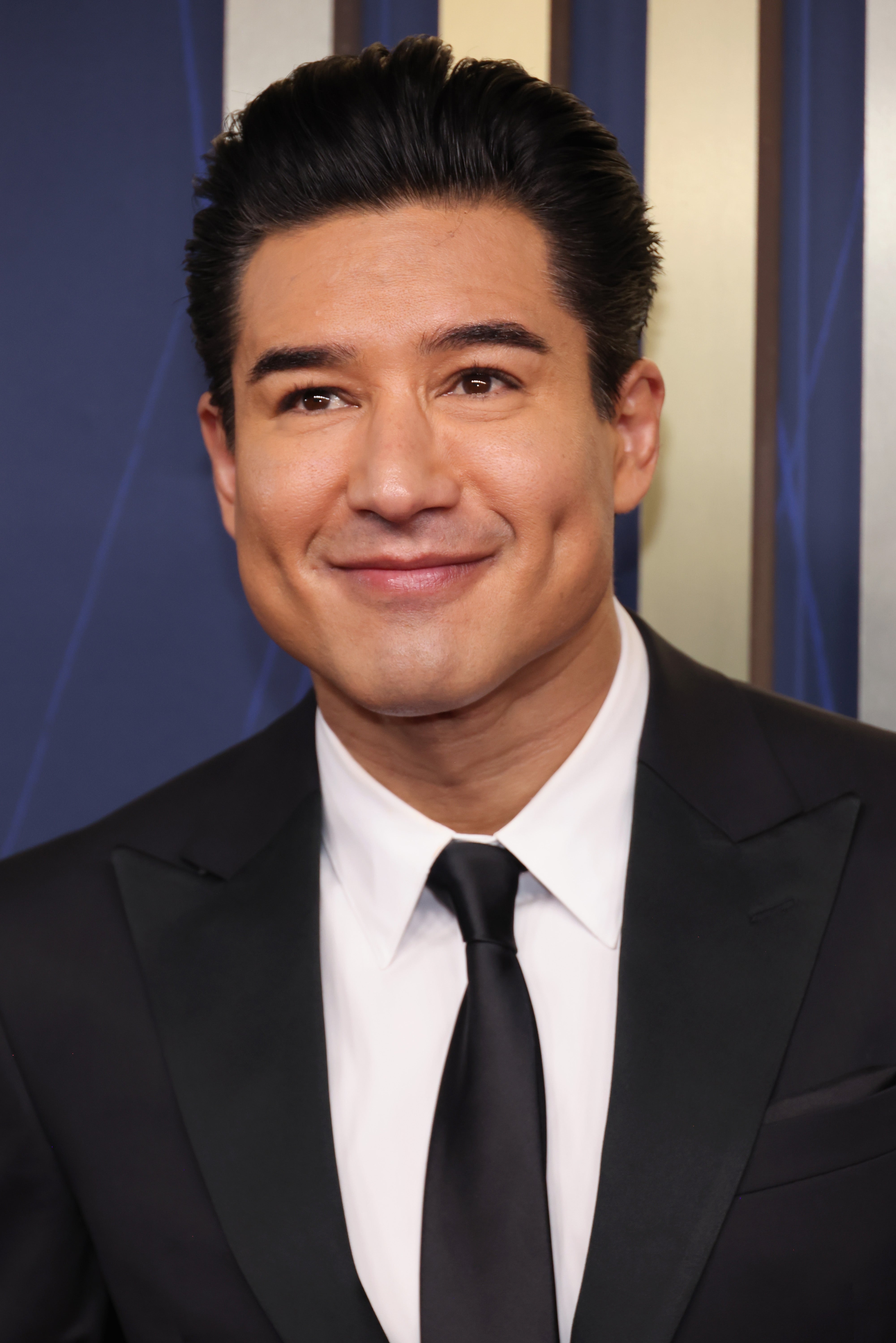 Mario Lopez smiles, wearing a black suit and tie