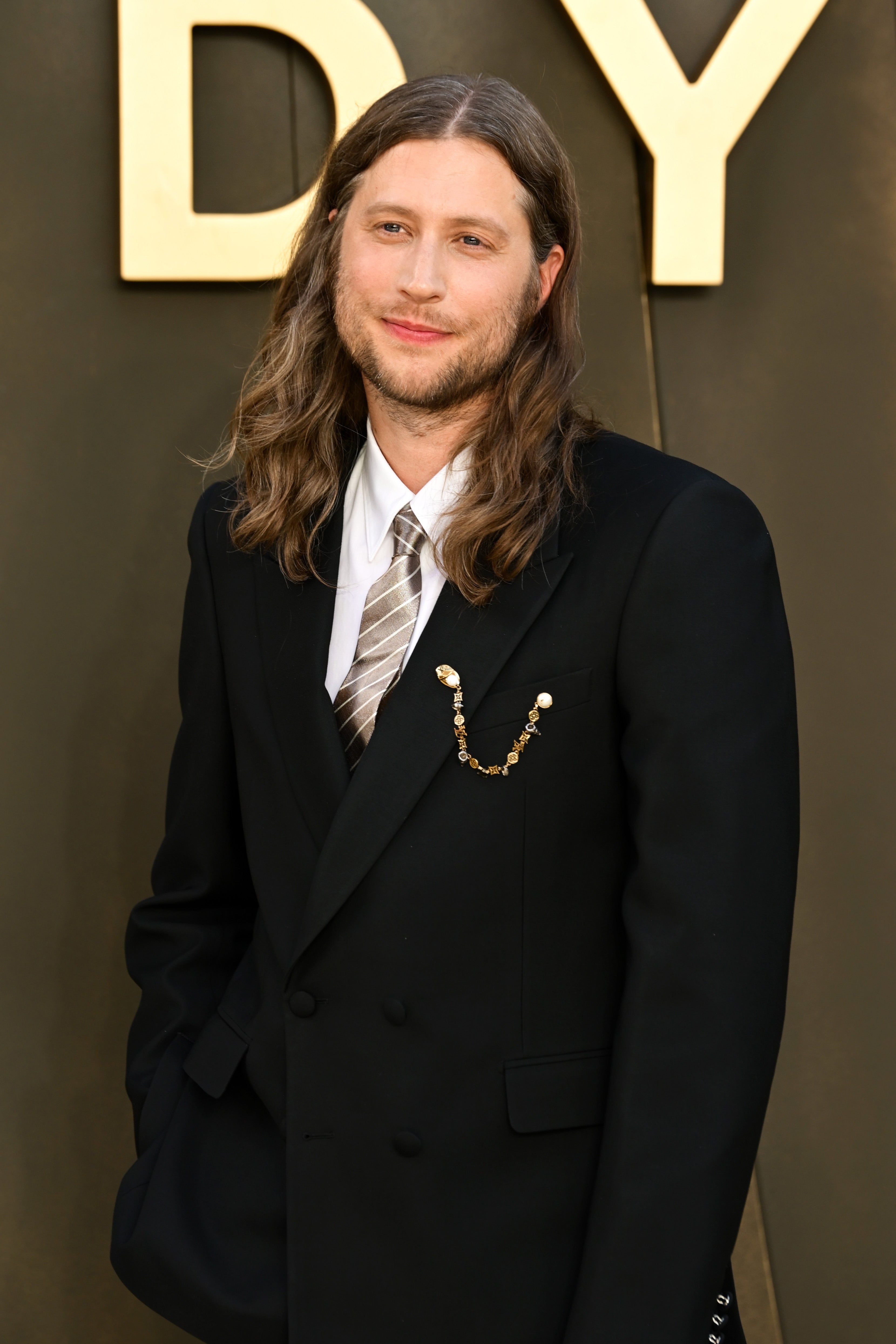 Person in a stylish suit with a striped tie on a red carpet. They have long hair and are facing the camera with a slight smile