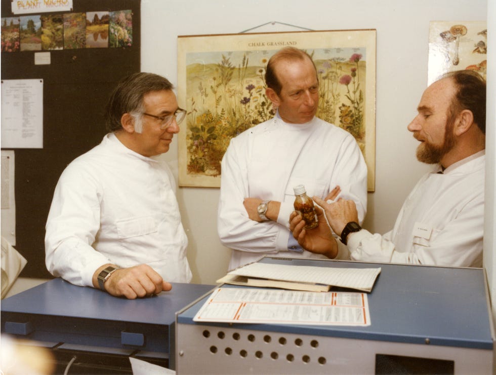 Three men in lab coats discuss a small bottle while standing in a laboratory environment with charts on the wall.