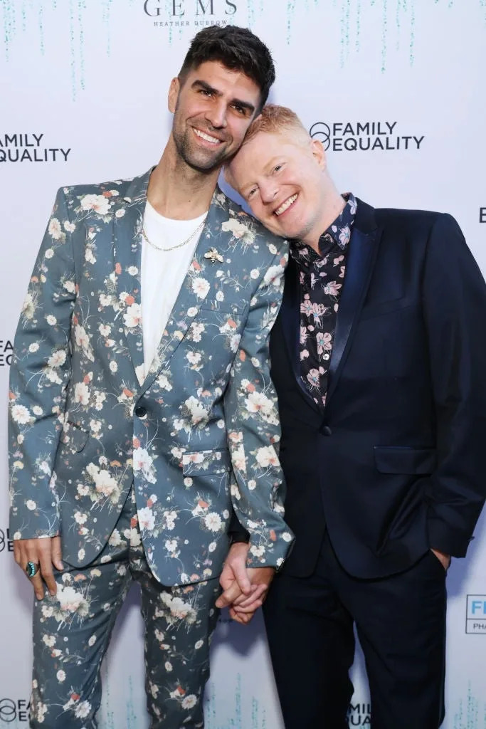 Two people holding hands on the red carpet. One in a floral suit, the other in a dark suit with a patterned shirt, attending an LGBT event