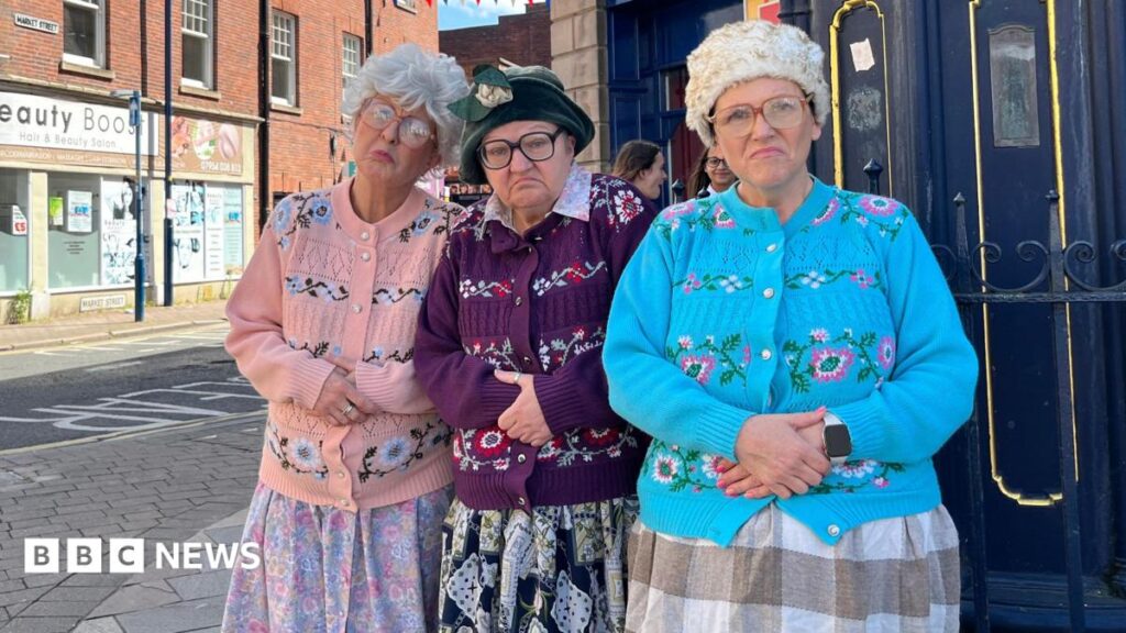 The Black Country grannies who want the world to dance Three women are standing together on a high street wearing colourful knitted cardigans with their hands folded on top of their stomach. They are all wearing glasses and have grey wigs on their heads.