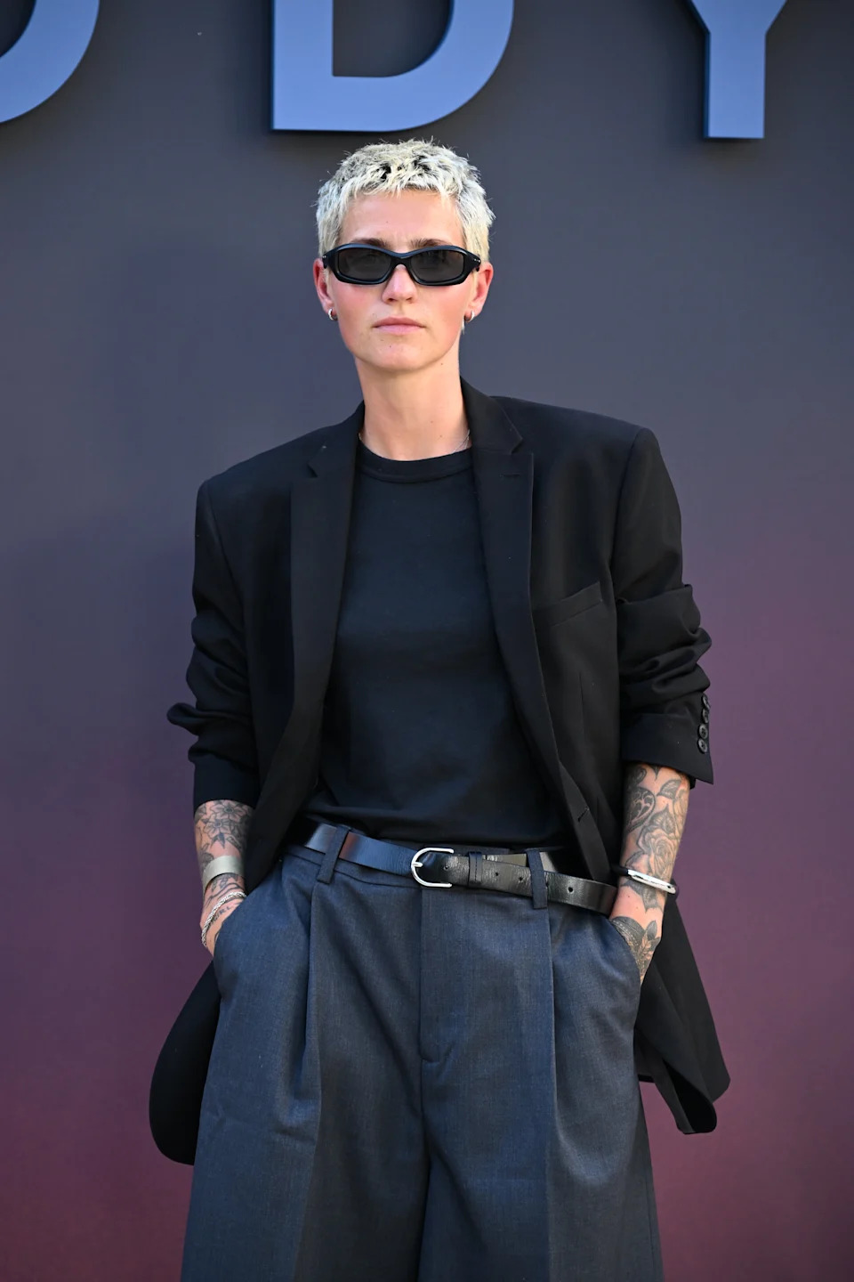 Person wearing a black blazer, dark shirt, sunglasses, and wide-leg trousers, posing confidently