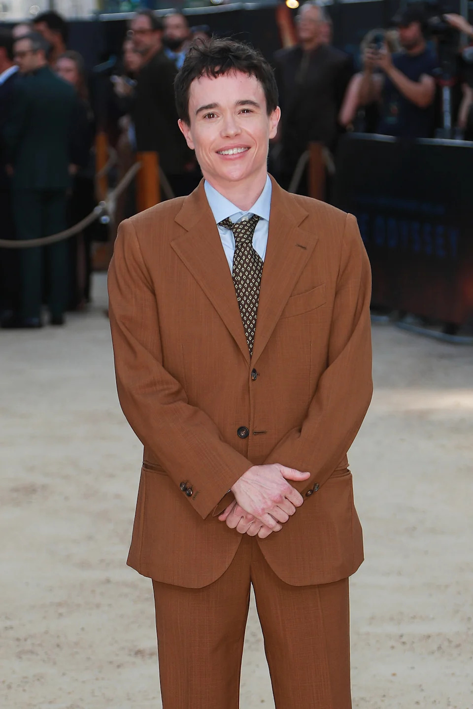 Person in a brown suit and tie smiling on a red carpet at a public event