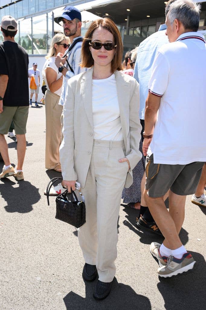 Claire Foy in a white suit and sunglasses, holding a black handbag, stands among a crowd.