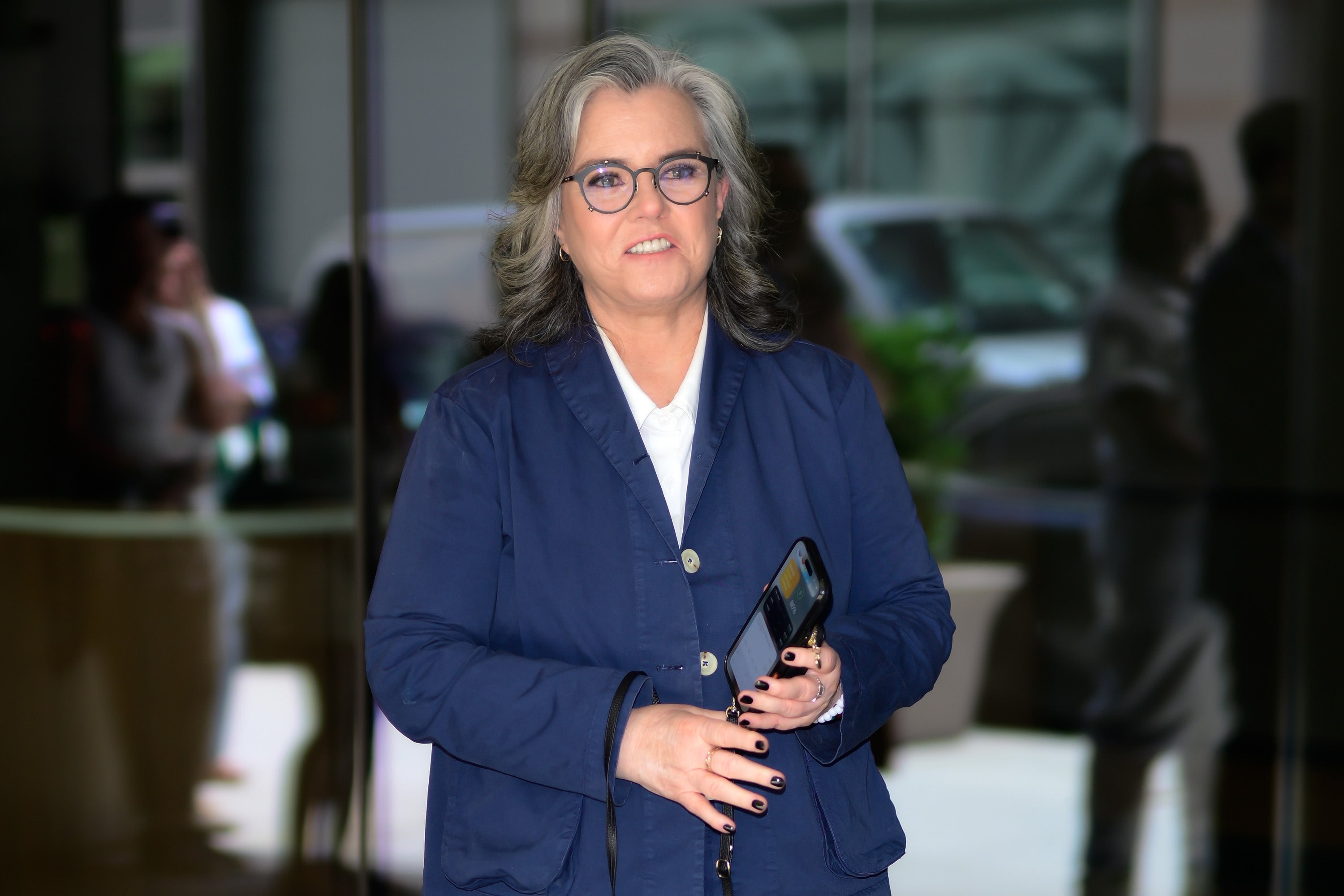 Rosie O'Donnell wearing a blue blazer and glasses, holding a phone, outside a building