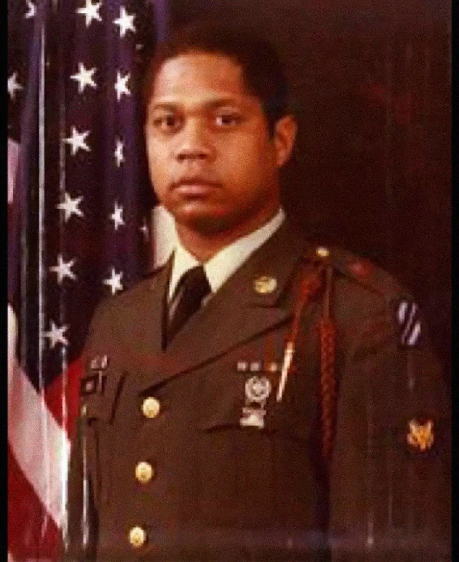 A man in a military uniform stands in front of an American flag, looking at the camera with a serious expression.