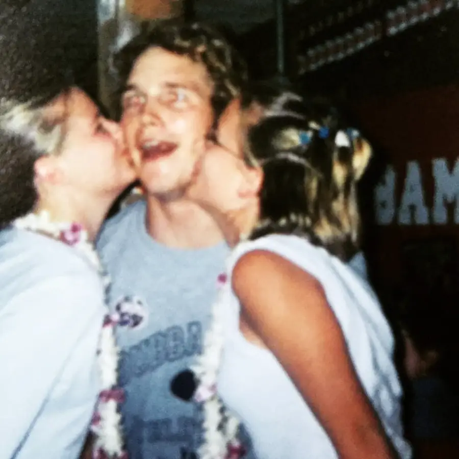 A young man smiles widely as two girls kiss him on the cheeks. All three wear leis and casual clothes, appearing to celebrate in a festive, joyful setting.