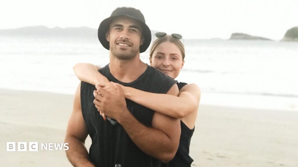 GriefTok: Woman says sharing loss of fiancé online ‘gives her purpose’ An image of a man and a woman standing on a beach. A blonde woman with sunglasses on her head has her arms around a man wearing a black vest and black bucket hat. They are both smiling at the camera.