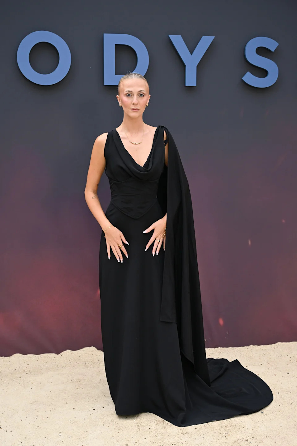 Person in elegant, flowing black gown with draped sash stands on sand against a backdrop with large letters "ODYS" visible