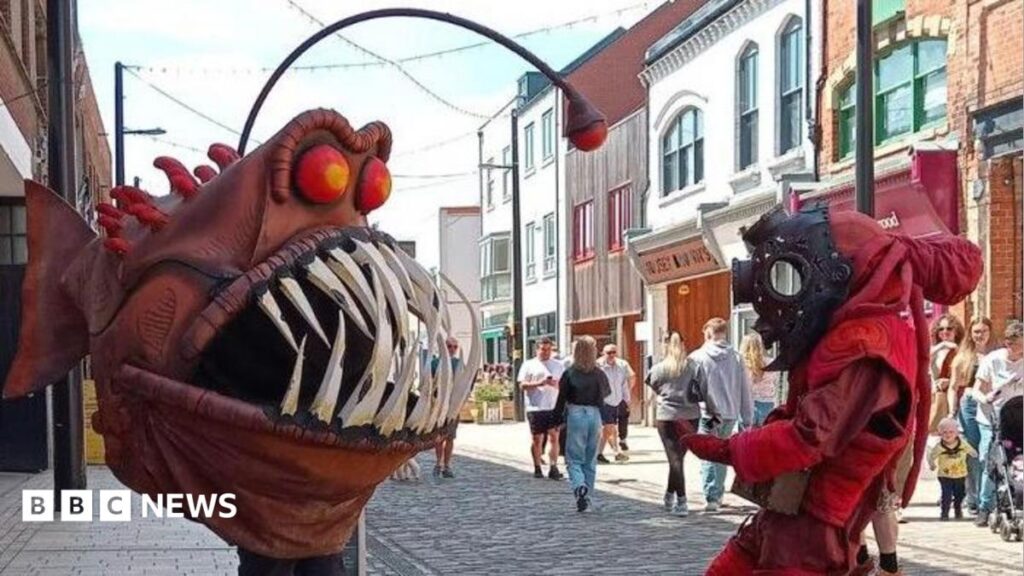 How fantastic beasts are hatched at a workshop in Hull The "Humber Monster" - a person wearing a large, brown angler fish costume with bulging red eyes and sharp white teeth, confronts a person wearing a red diving suit and black diving helmet, while standing in a pedestrian high street as people watch.