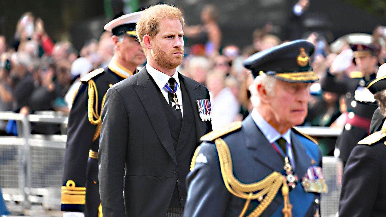 King Charles walking in a blue uniform looking somber in front of Prince Harry in a dark suit.
