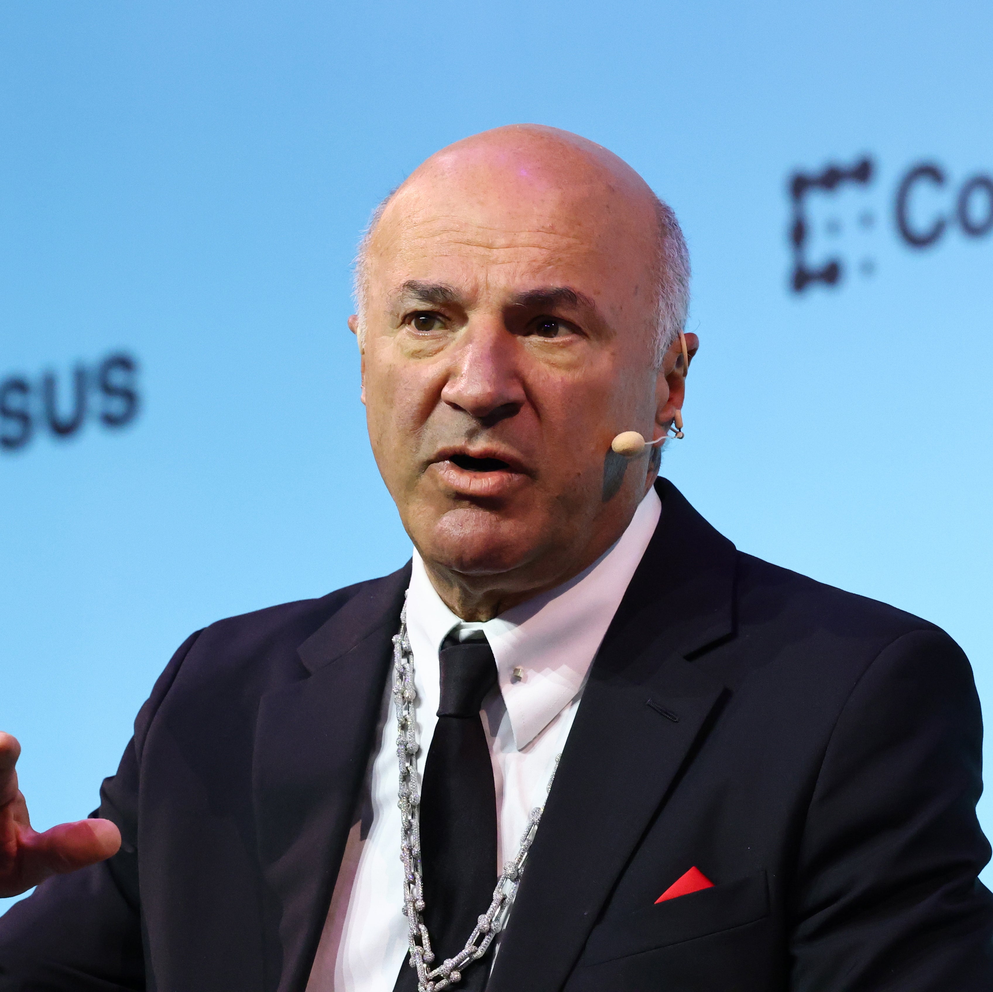 Kevin O’Leary in a suit with a microphone attached speaks at a conference panel, gesturing as they talk