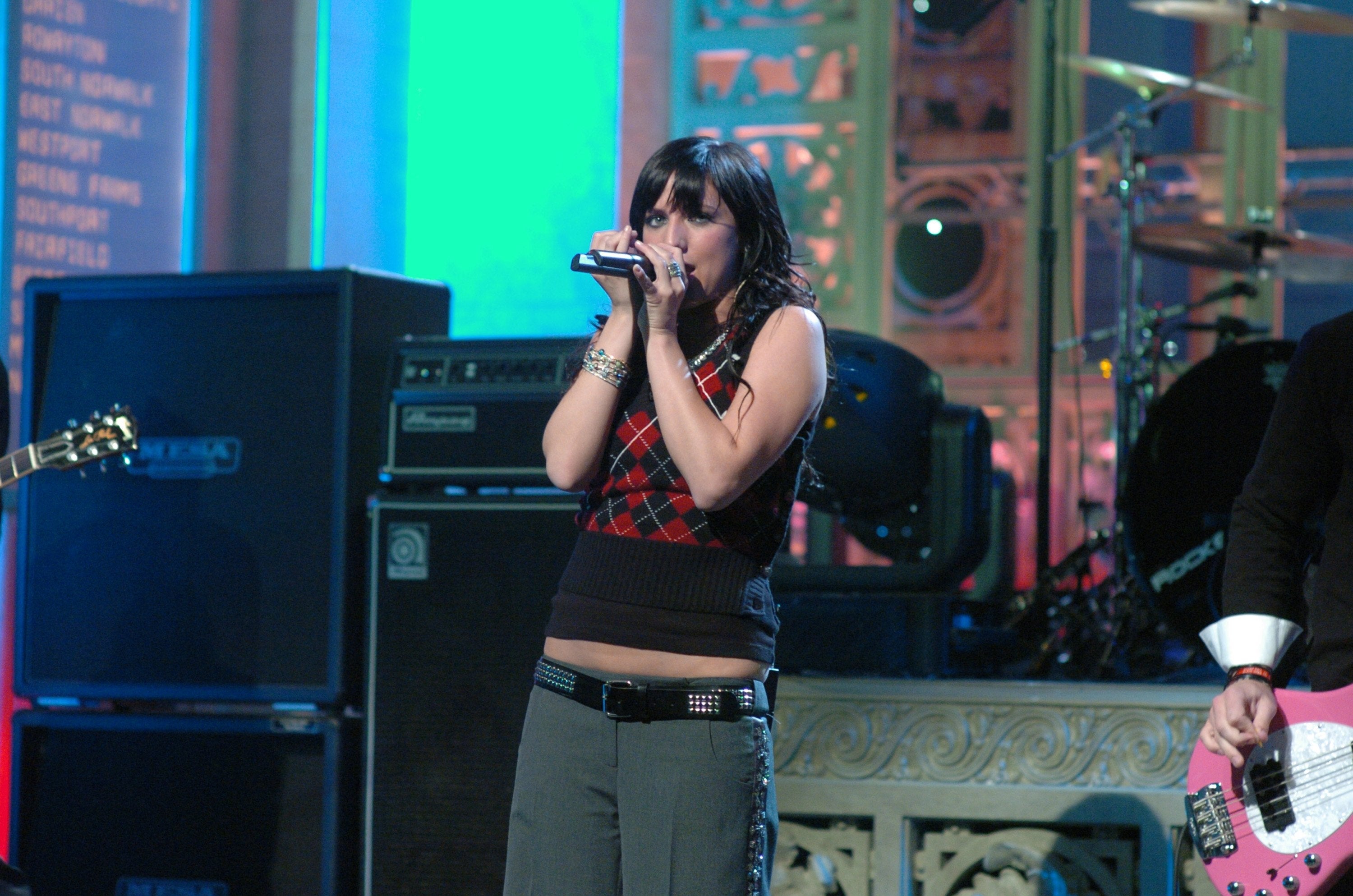 Ashlee Simpson in sleeveless plaid top singing into a microphone on a stage with band equipment in the background