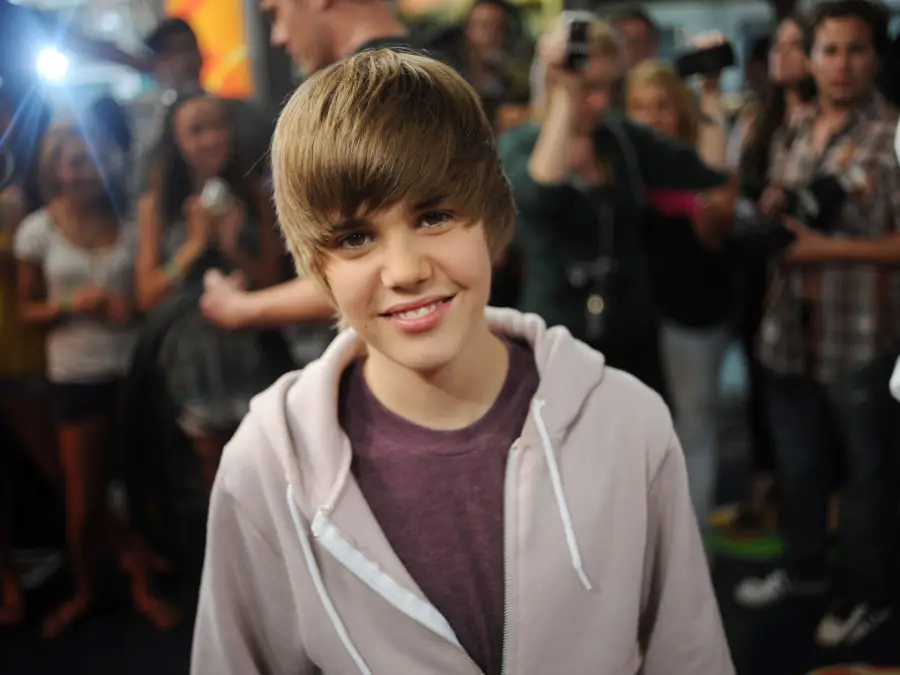 A young person with side-swept brown hair smiles at the camera, wearing a light hoodie over a maroon shirt. A crowd of people, some with cameras, can be seen in the blurred background.
