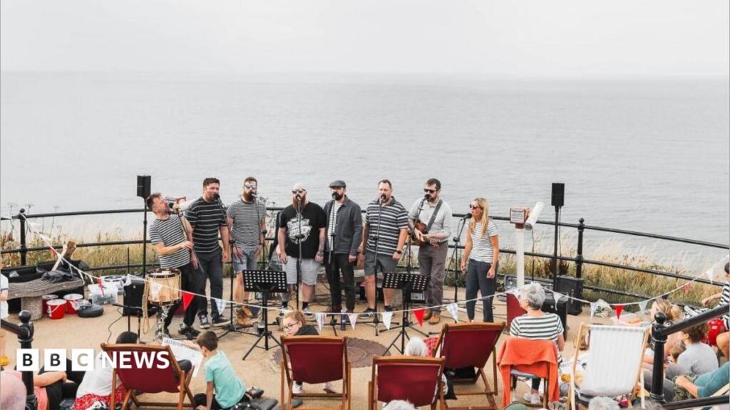 Eight performers stand in a line on the promenade and sing into microphones. There is a crowd of people on the stairs and in deck chairs watching on. the sea behind them is grey and calm, and a similar colour to the sky above.